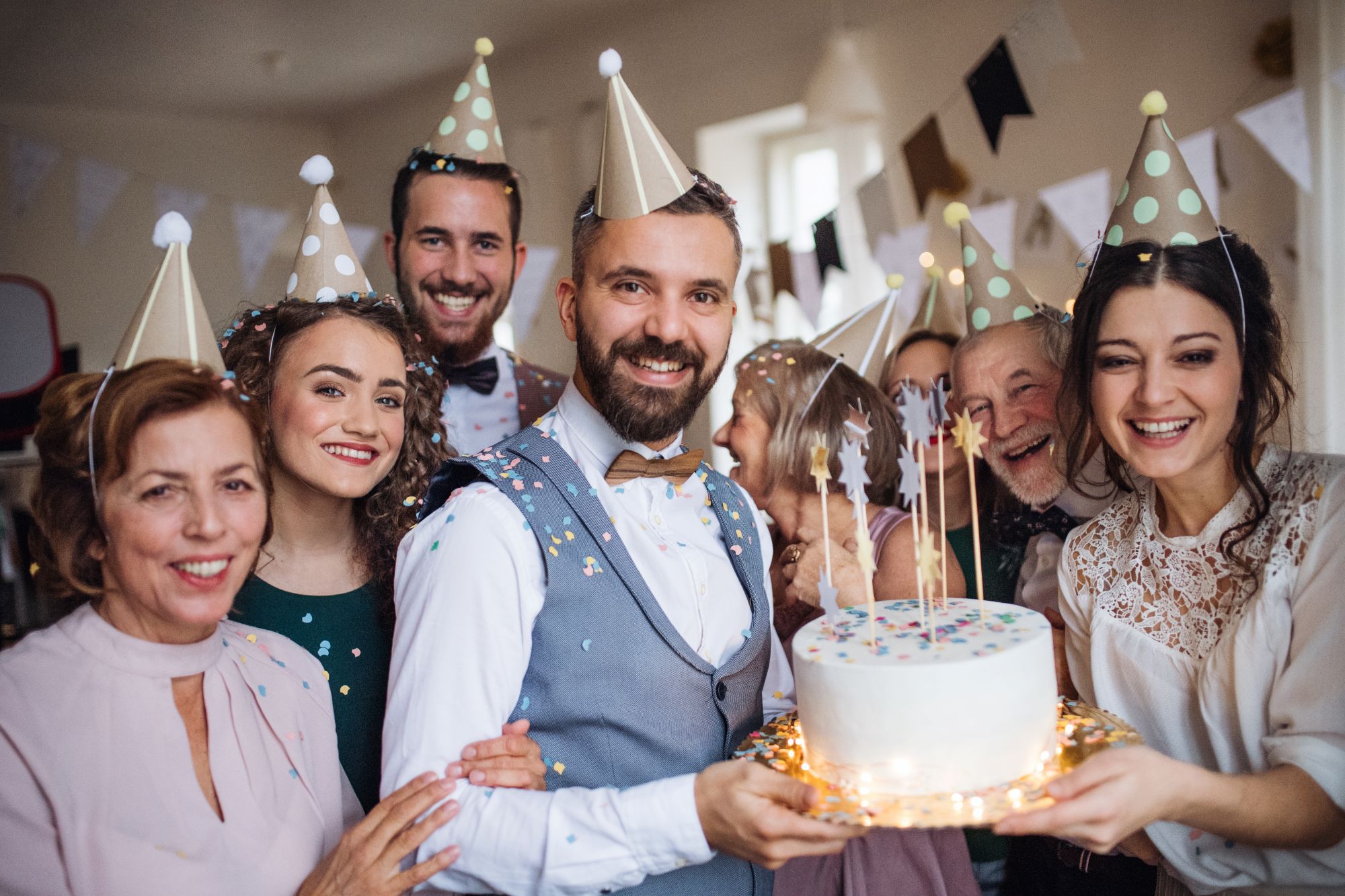 Multi-generational family in party hats smiling around a birthday cake with sparklers