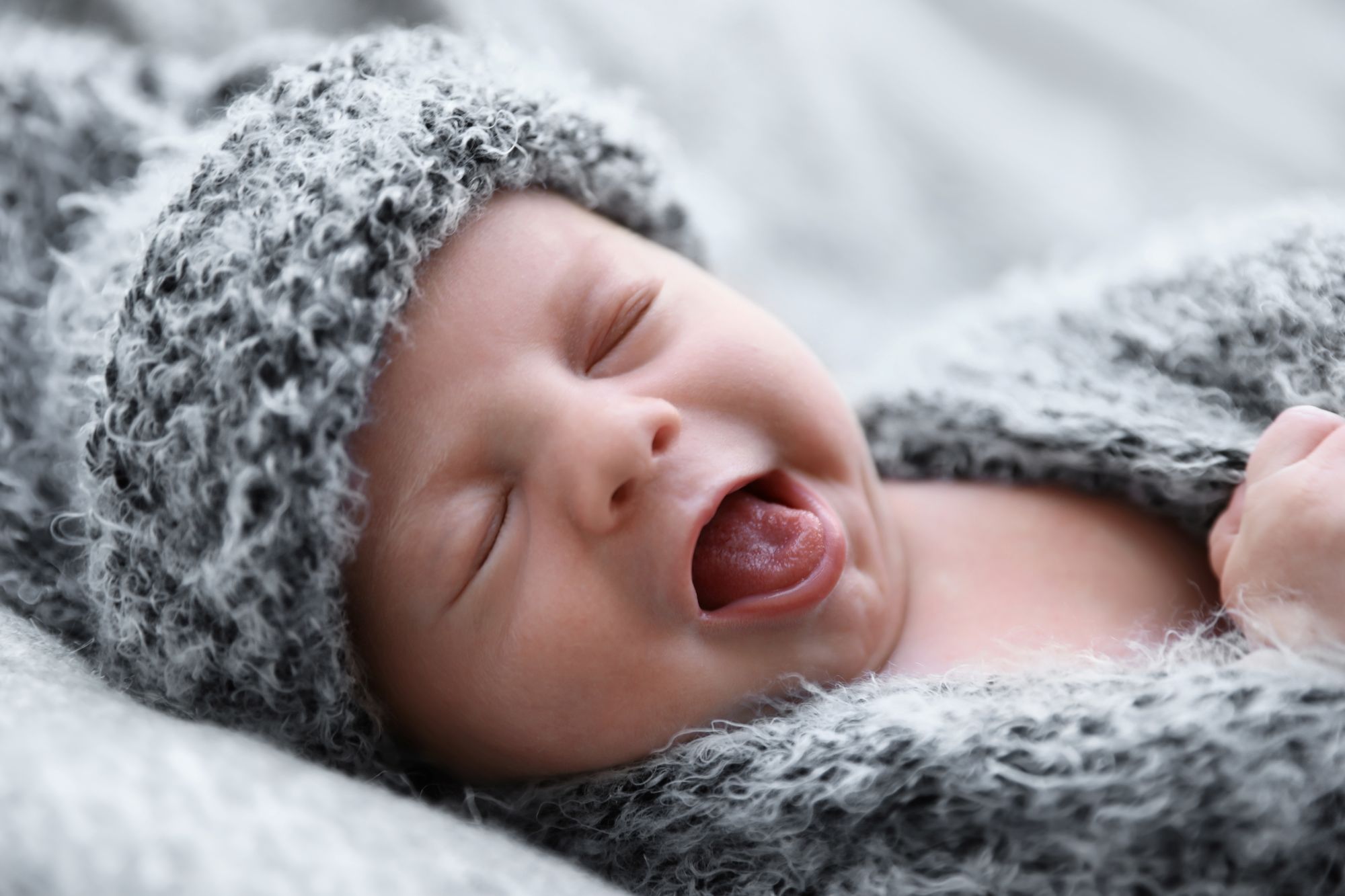 Sleepy newborn yawning while swaddled in a cozy grey knit wrap
