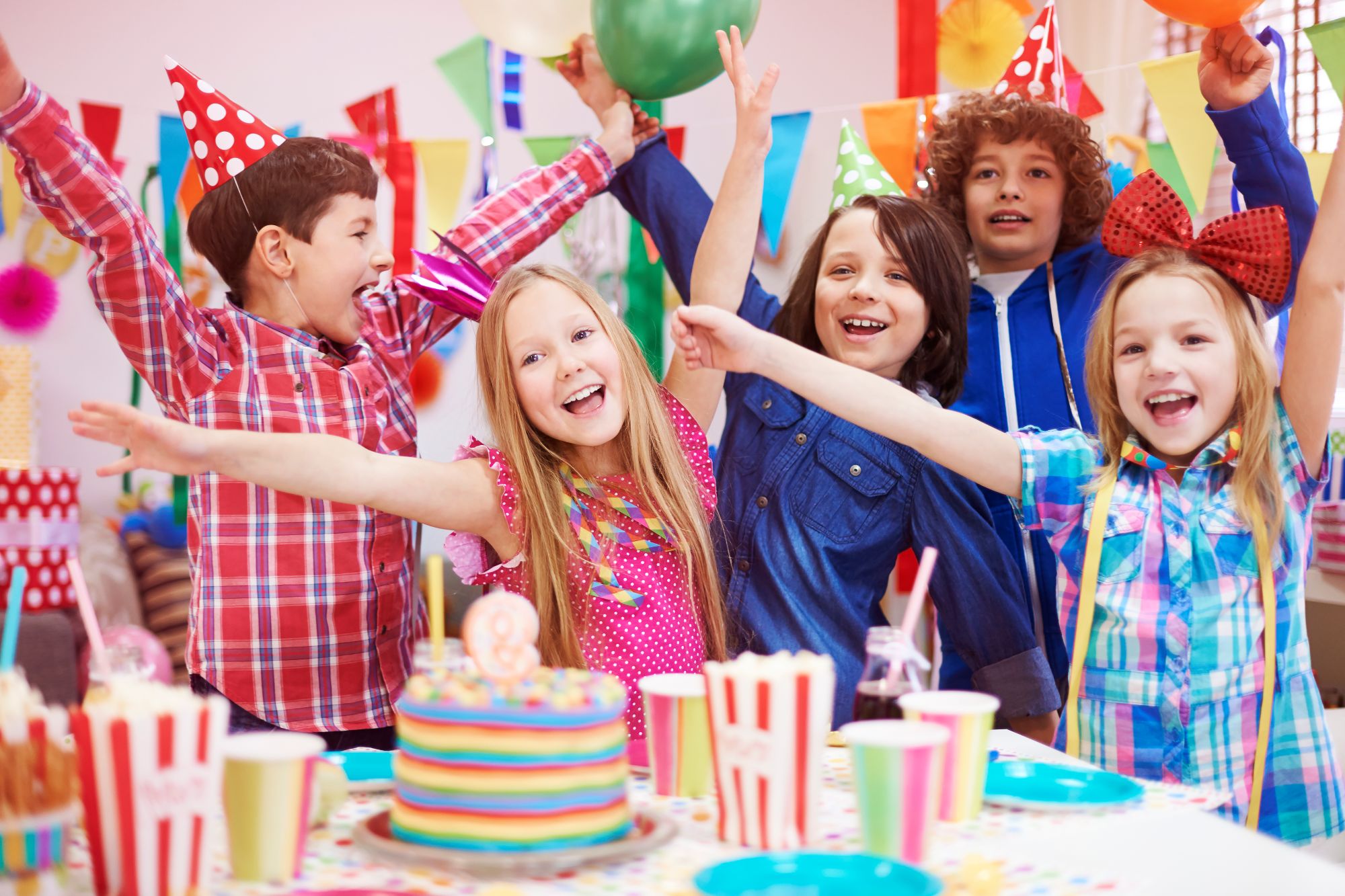Group of children in party hats cheering at a colourful birthday party with cake