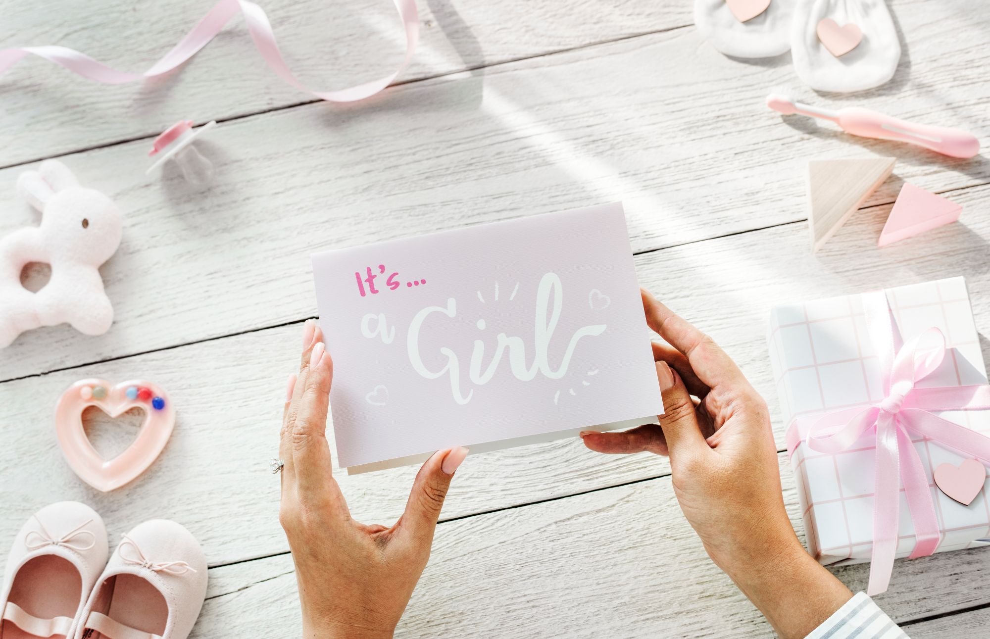 Hands holding an Its a Girl pink baby shower card with bunny, shoes and gift