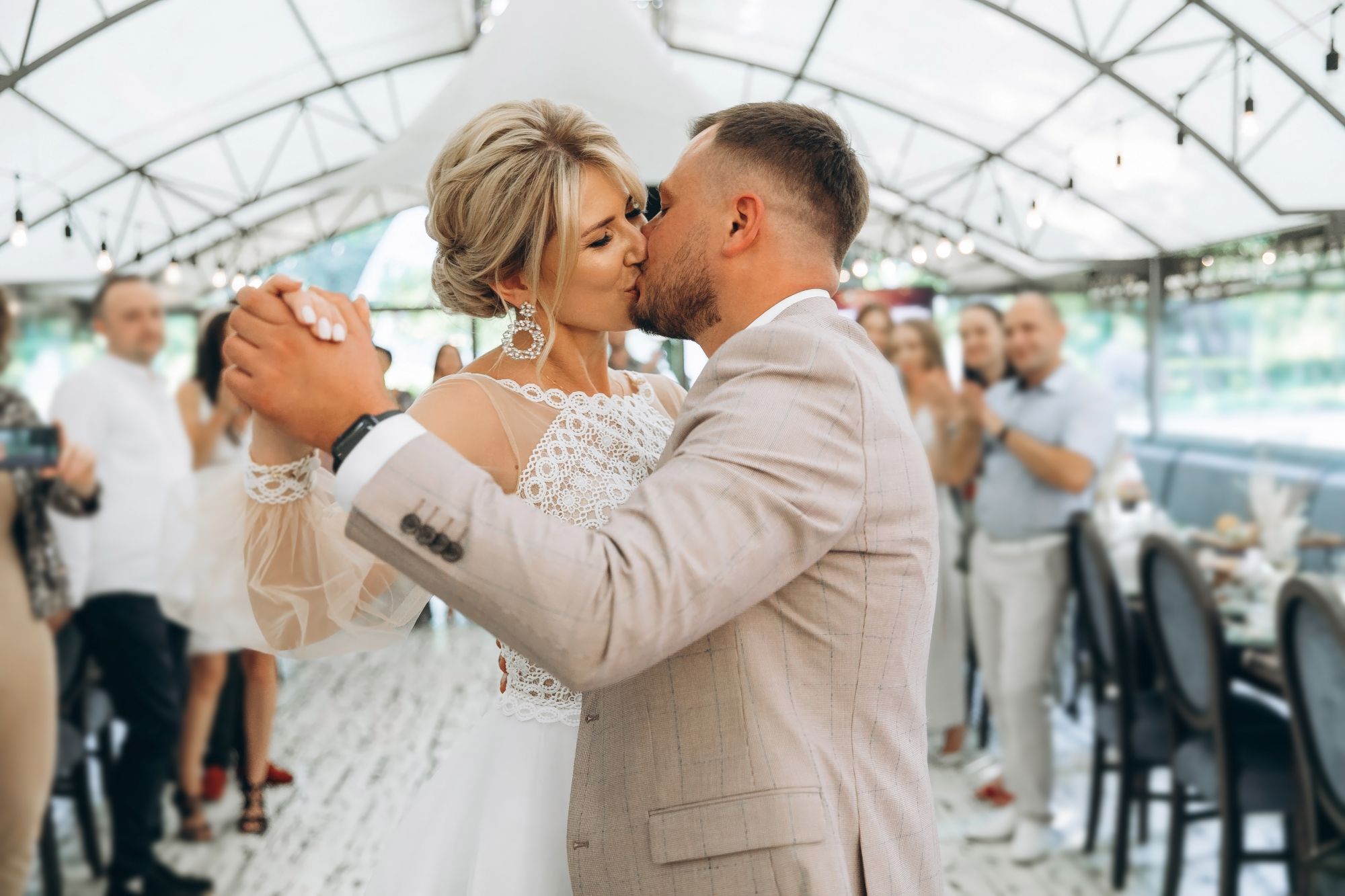 Newlyweds kissing during their first dance under string lights with guests watching