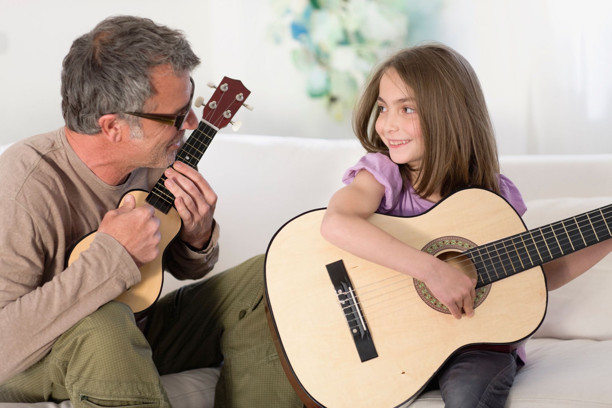 Father playing ukulele while his daughter plays acoustic guitar on the couch
