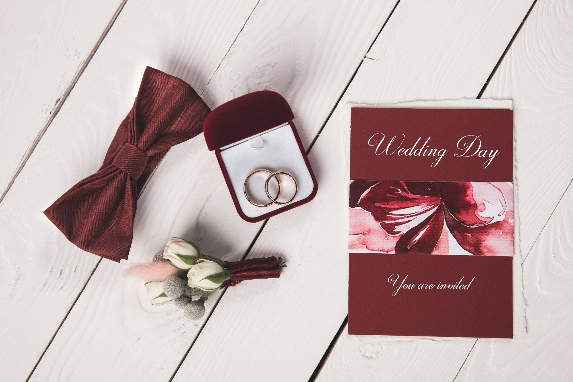 Burgundy wedding day invitation card with rings and bow tie on white wood
