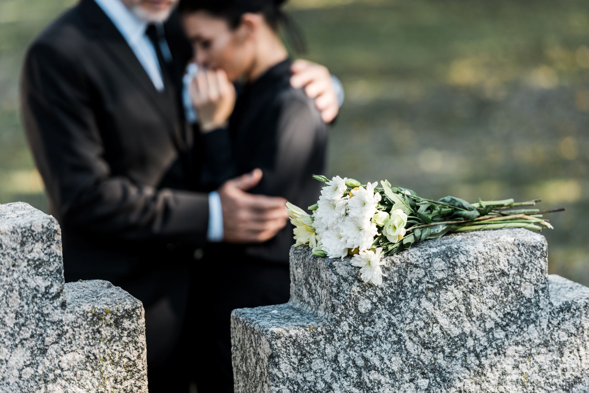 Grieving couple in black embracing beside a granite memorial with white flowers