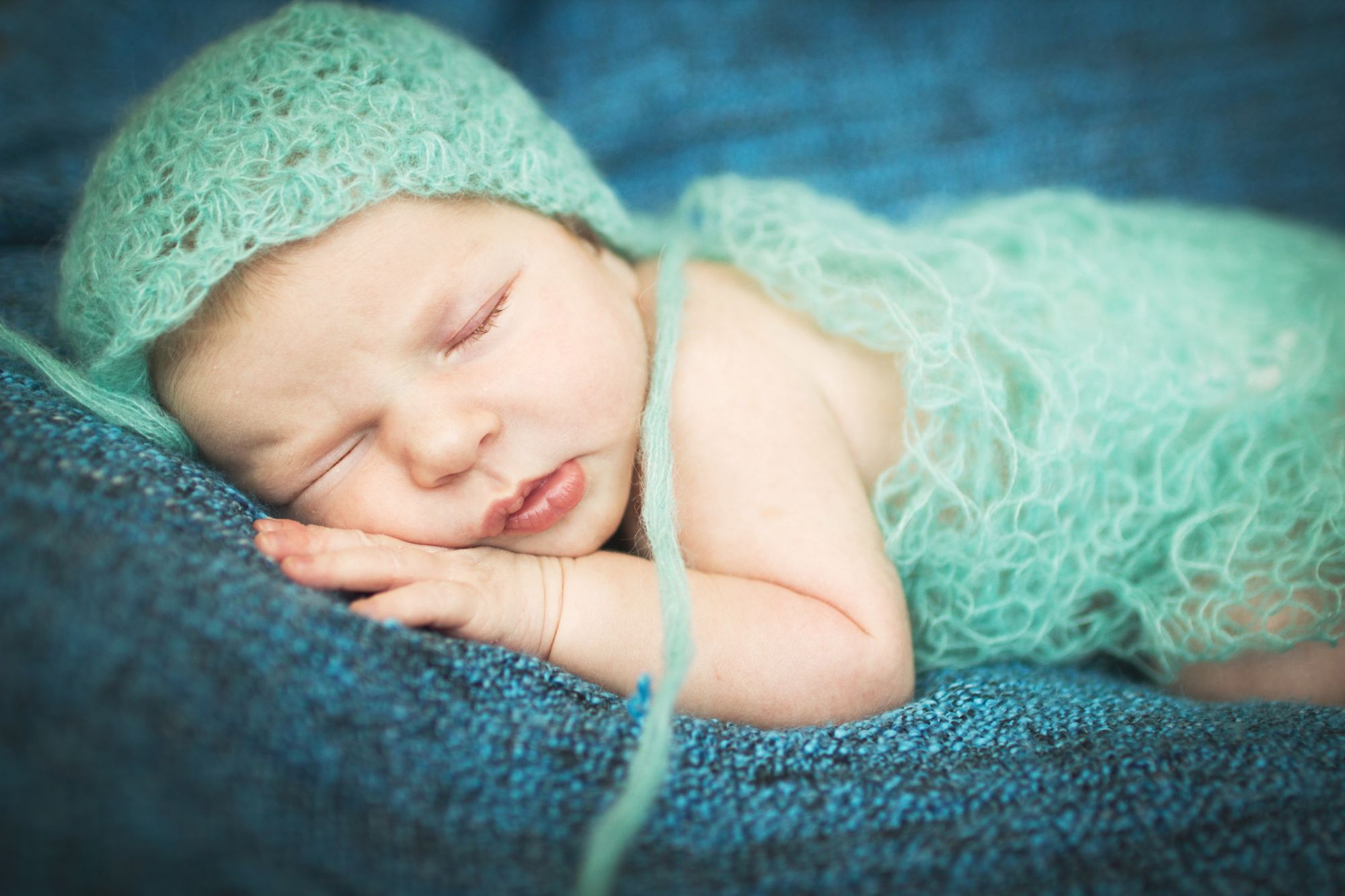 Newborn baby peacefully sleeping in a soft mint knit bonnet and wrap on a blue blanket