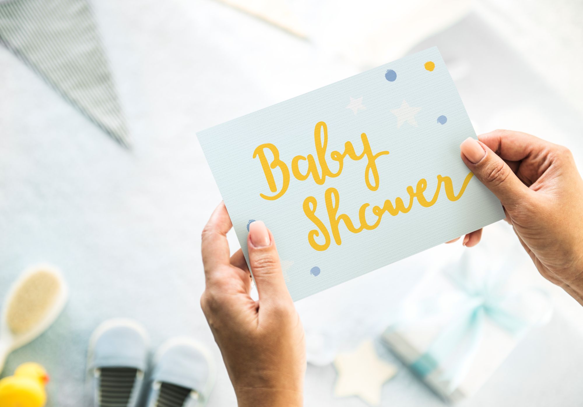 Hands holding a pastel blue Baby Shower greeting card surrounded by baby items